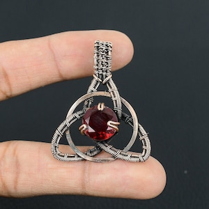 May include: A copper wire wrapped pendant with a red gemstone in the center. The pendant is in the shape of a Celtic knot.