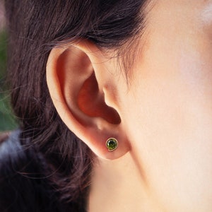 May include: A close-up of a person's ear with a small green gemstone earring. The earring is set in gold and has a round shape.