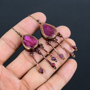 Puede incluir: Pendientes colgantes de color cobre con piedras fucsia grandes e irregulares. Cada pendiente tiene una piedra en forma de lágrima en la parte superior, con tres varillas de cobre colgantes, cada una terminada en pequeñas piedras fucsia redondas.