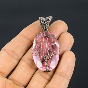 Puede incluir: Un colgante de piedra preciosa rosa con un dise&ntilde;o de &aacute;rbol envuelto en alambre. La piedra ovalada es de color rosa claro, y el &aacute;rbol est&aacute; hecho de alambre color cobre. La fianza est&aacute; hecha de alambre m&aacute;s oscuro y apretado. El colgante est&aacute; sobre un fondo oscuro.