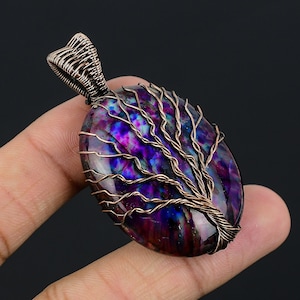 Puede incluir: Un colgante envuelto en alambre de cobre con un diseño de árbol de la vida sobre una piedra preciosa iridiscente púrpura y azul.