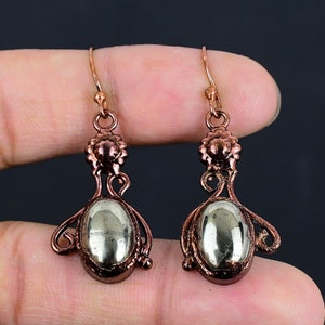 May include: A pair of copper dangle earrings featuring oval-shaped pyrite stones. The earrings have a delicate, ornate design with a floral motif.