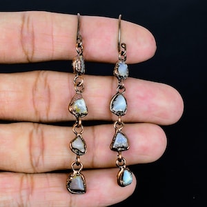 May include: Copper-toned dangle earrings with four irregularly shaped, light-colored stones. The stones have a pearlescent sheen and are connected by small copper links. The earrings are displayed against a dark background.