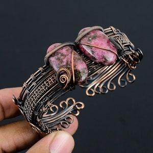 May include: A handcrafted copper wire cuff bracelet featuring two pink and green gemstones. The bracelet has intricate wirework with a woven design and decorative swirls. The gemstones are secured with wire wrapping, creating a unique and artistic piece of jewelry.