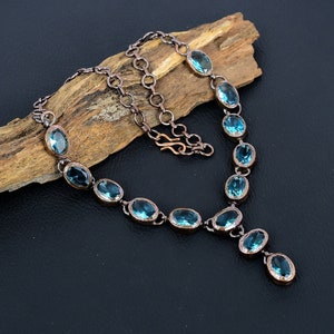 May include: A copper necklace with a teardrop pendant features oval-shaped blue gemstones. The necklace is displayed on a piece of driftwood.