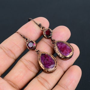 Op de afbeelding: Koperkleurige oorbellen met elk drie robijnrode edelstenen. De oorbellen hebben een druppelvormige steen, een ronde steen en een vierkante steen. De edelstenen zijn gezet in een getextureerde koperen lijst.