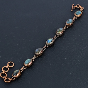 Puede incluir: Un brazalete de cobre con un diseño de cadena de eslabones y piedras preciosas de labradorita de forma ovalada. El brazalete tiene un cierre de langosta.