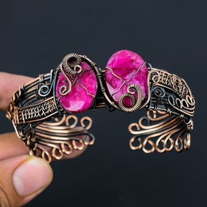 May include: A handcrafted copper cuff bracelet with two oval, vibrant pink gemstones. The gemstones are intricately wire-wrapped, and the bracelet has an open design with detailed metalwork. The bracelet's colour palette includes copper, black, and pink.
