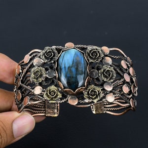 May include: A copper and brass cuff bracelet with a large oval labradorite gemstone surrounded by floral designs. The bracelet has a woven wire design and is adjustable.