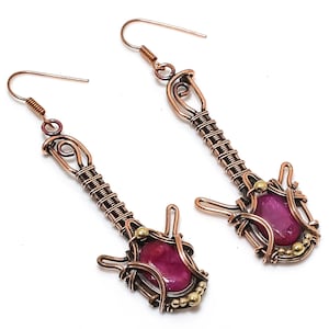 May include: A pair of handcrafted copper wire earrings. Each earring features a guitar-shaped design with a vibrant magenta gemstone at the base, accented with small gold-colored beads. The earrings have a hook closure.