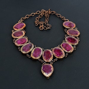 May include: A copper necklace with a pendant and a row of oval-shaped, deep pink gemstones. The gemstones are set in copper.