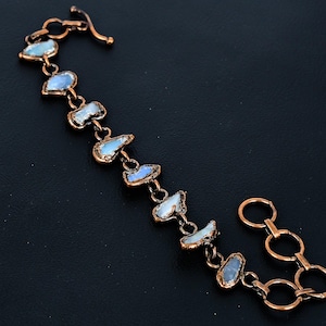 以下が含まれることがあります： 不規則な形をした、水色と白の石が連なった銅色のブレスレット。石は小さな銅製のコネクタと留め金で繋がれています。ブレスレットは黒い背景に展示されています。