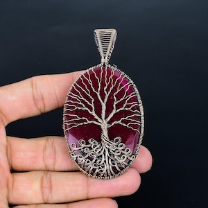 May include: An oval-shaped pendant featuring a deep magenta stone base wrapped in dark copper wire. The wire is intricately coiled to form the shape of a tree with branches extending upward and roots curling at the bottom, topped with a wire bail.