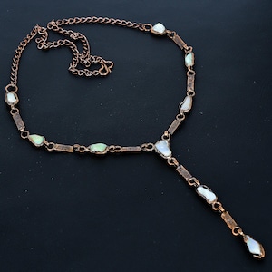 Puede incluir: Un collar de tono cobrizo con un colgante en forma de Y. El collar presenta pequeñas piedras preciosas irregulares, blanquecinas, engastadas en eslabones rectangulares y de cadena. Las piedras preciosas tienen una sutil calidad iridiscente.