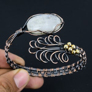 May include: A wire-wrapped cuff bracelet featuring a large, oval, iridescent white moonstone cabochon. The band is crafted from dark, oxidized copper wire with intricate woven patterns and a decorative feather-like motif accented with small gold-toned beads.
