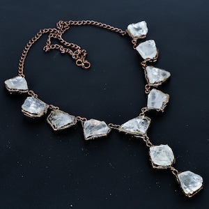 Puede incluir: Un collar de color cobre con piedras de cuarzo transparente de forma irregular. Las piedras están engastadas en metal color cobre y conectadas por una delicada cadena. El collar tiene un diseño de colgante en forma de Y.