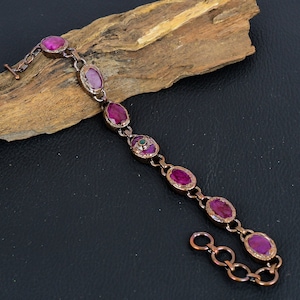 May include: A copper-toned bracelet featuring oval-shaped, deep pink gemstones. The bracelet has a toggle clasp and is displayed on a piece of wood against a dark background. The gemstones are set in a copper-colored metal.