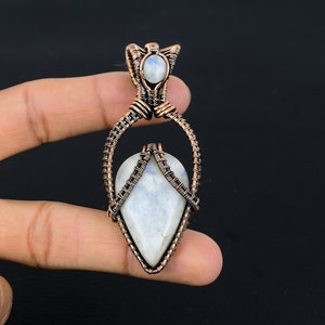 May include: A teardrop-shaped moonstone pendant wrapped in copper wire. The pendant is held by a finger.