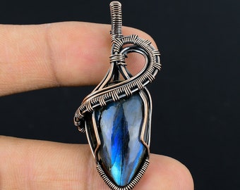 Può includere: Un ciondolo fatto a mano con una pietra di labradorite a forma di goccia, avvolta in modo intricato con filo di rame. La pietra mostra sfumature iridescenti blu e verdi. La filigrana forma un'ansa decorativa per una collana.