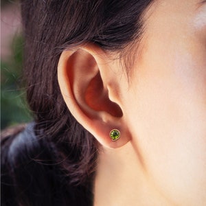 May include: A close-up of a person's ear with a small green gemstone earring. The earring is set in gold and has a round shape.