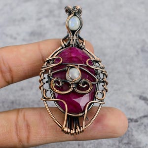 May include: A copper wire wrapped pendant featuring a large, faceted, pink ruby gemstone, a smaller white pearl, and a smaller, iridescent moonstone. The pendant is oval shaped and has a delicate, intricate design.