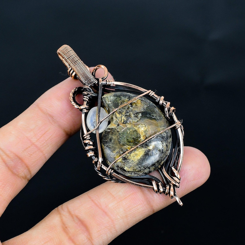 Puede incluir: Colgante artesanal con un amonita fosilizada encapsulada en un marco de alambre de cobre. El f&oacute;sil muestra tonos de verde, marr&oacute;n y dorado. El trabajo de alambre est&aacute; intrincadamente dise&ntilde;ado, creando una pieza de joyer&iacute;a &uacute;nica y art&iacute;stica.