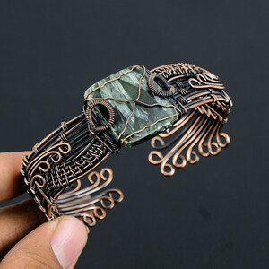 May include: A copper wire wrapped bracelet with a green and black stone set in the center. The bracelet is made with a woven wire design and has a decorative, curled wire design on the sides.
