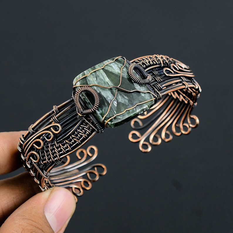 May include: A copper wire wrapped bracelet with a green and black stone set in the center. The bracelet is made with a woven wire design and has a decorative, curled wire design on the sides.