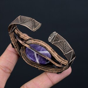 May include: Copper wire cuff bracelet with a central oval amethyst stone. The amethyst is a deep purple with white veining. The bracelet's wirework has a woven pattern. The bracelet is open at the ends.