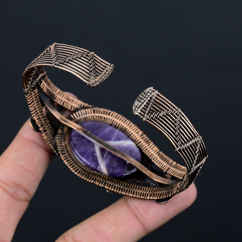 May include: Copper wire cuff bracelet with a central oval amethyst stone. The amethyst is a deep purple with white veining. The bracelet's wirework has a woven pattern. The bracelet is open at the ends.