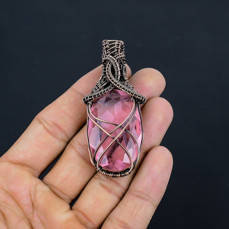 May include: A pink gemstone pendant wrapped in copper wire. The pendant is oval shaped and has a faceted design. The wire is wrapped around the stone in a decorative pattern.