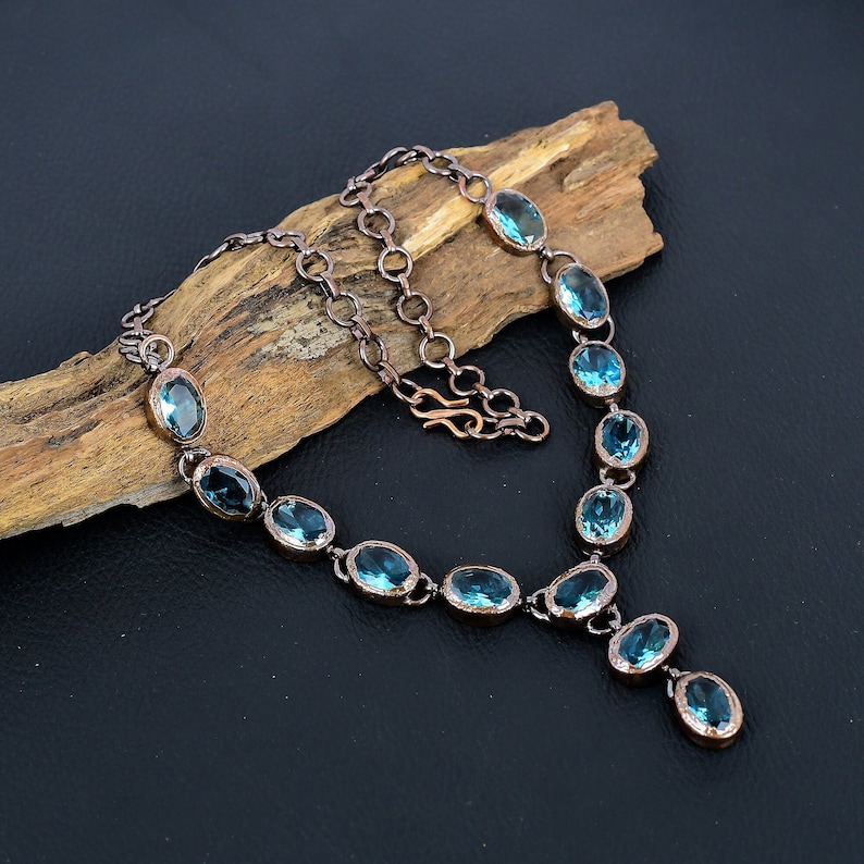 May include: A copper necklace with a teardrop pendant features oval-shaped blue gemstones. The necklace is displayed on a piece of driftwood.