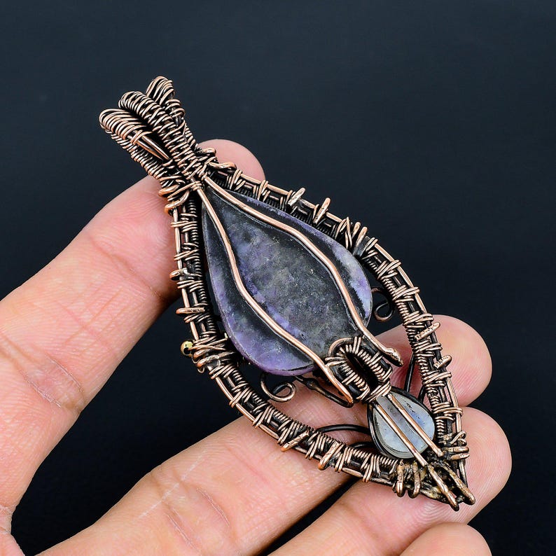May include: A handcrafted copper wire pendant with a teardrop-shaped purple stone and a smaller, light-colored stone at the bottom. The pendant is intricately woven with copper wire, creating a unique, artistic design. The pendant is approximately 2 inches long.
