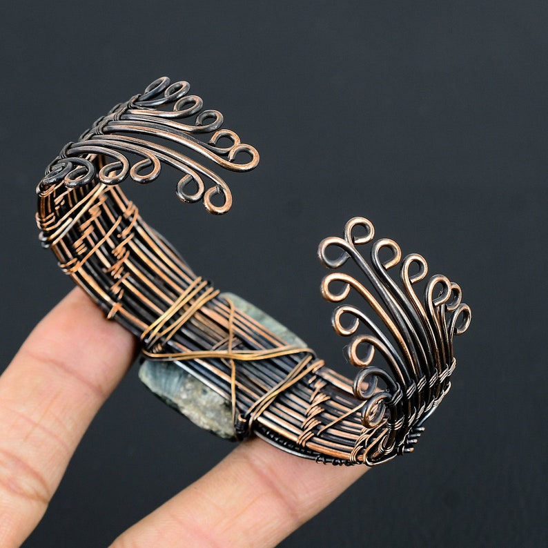 May include: A copper wire bracelet with a green stone set in the center. The bracelet is decorated with intricate wirework and has a delicate, flowing design.