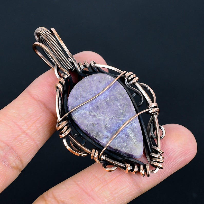Peut inclure: Un pendentif artisanal avec une pierre en forme de goutte, de couleur lavande. La pierre est sertie dans un cadre en fil de cuivre avec un tissage complexe et un support en m&eacute;tal fonc&eacute;. Le pendentif est con&ccedil;u pour &ecirc;tre port&eacute; en collier.