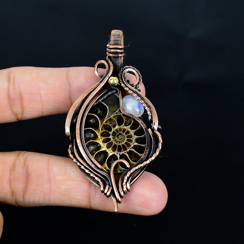 May include: A handcrafted copper pendant featuring an ammonite fossil with a spiral pattern, accented by a small moonstone and a gold bead. The pendant is intricately wired and has a dark background.