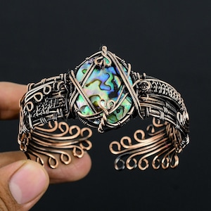 May include: Copper wire cuff bracelet with an abalone shell centerpiece. The bracelet features intricate wire wrapping and a natural, iridescent stone. The open design allows for easy wear and showcases the handcrafted details.