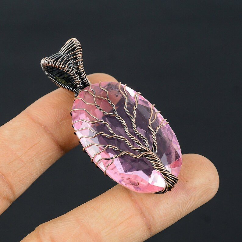 Puede incluir: Un colgante de piedra preciosa rosa con un dise&ntilde;o de &aacute;rbol de la vida. La piedra rosa ovalada y facetada est&aacute; encerrada en alambre color cobre, formando las ramas y el tronco del &aacute;rbol. La fianza es un dise&ntilde;o de alambre oscuro y retorcido.
