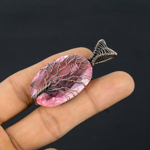 Puede incluir: Un colgante de piedra preciosa rosa con un dise&ntilde;o de &aacute;rbol de la vida. El colgante presenta una piedra rosa facetada de forma ovalada, envuelta en alambre de cobre, con el alambre formando la forma de un &aacute;rbol. La fianza tambi&eacute;n es de alambre de cobre.