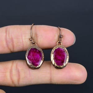 May include: A pair of oval-shaped ruby earrings. Each earring features a vibrant, deep pink ruby stone set in a gold-colored, textured frame. The earrings have a simple hook design for pierced ears, showcasing a classic jewelry style.