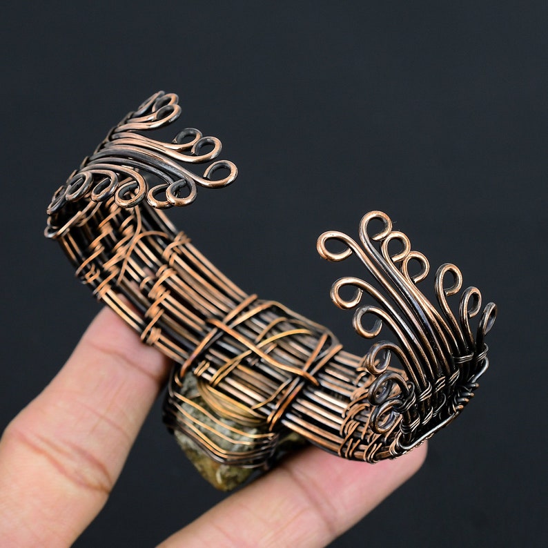 May include: A copper wire bracelet with a woven design and a green stone accent. The bracelet has a decorative, swirling design on the ends.