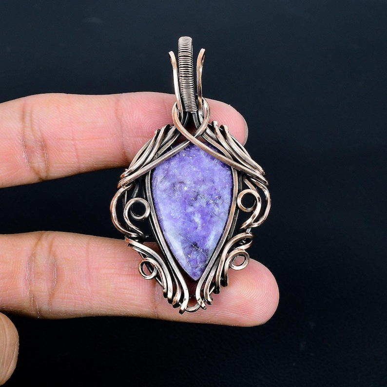 Peut inclure: Pendentif artisanal avec une pierre violette mouchet&eacute;e en forme de goutte d'eau. La pierre est sertie dans un design complexe de fil de cuivre avec des motifs tourbillonnants et une boucle pour une cha&icirc;ne. Le pendentif est tenu sur un fond sombre.