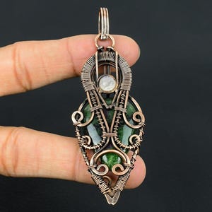 May include: A handcrafted copper wire pendant with a green stone centre and a smaller white stone. The pendant has intricate wire wrapping and a loop for a chain.