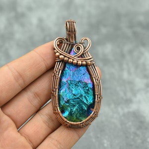 Puede incluir: Un colgante envuelto en alambre de cobre con una piedra preciosa azul y verde iridiscente. El colgante tiene forma de lágrima y presenta un diseño de alambre tejido.