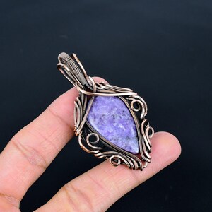Peut inclure: Un pendentif artisanal avec une pierre triangulaire de couleur lavande sertie dans un cadre en fil de cuivre. Le filigrane pr&eacute;sente des volutes et des boucles complexes, cr&eacute;ant une bordure d&eacute;corative autour de la pierre. Le pendentif est con&ccedil;u pour &ecirc;tre port&eacute; comme bijou.