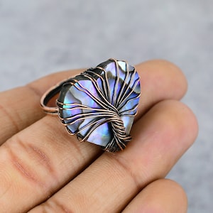 May include: A copper-colored ring featuring a heart-shaped abalone stone. The iridescent stone is wrapped with copper wire in a tree-like design. The ring is held in a hand against a neutral background.