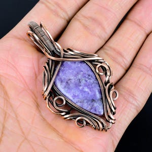 Peut inclure: Un pendentif artisanal avec une pierre violette polie de forme triangulaire, sertie dans une monture en fil de cuivre. Le filigrane pr&eacute;sente des volutes et des boucles complexes, cr&eacute;ant un design orn&eacute;. Le pendentif est con&ccedil;u pour &ecirc;tre port&eacute; en collier.