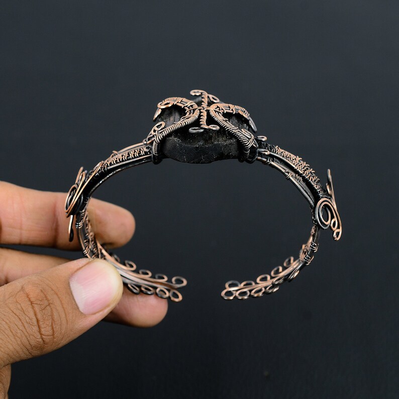 May include: A copper wire bracelet with a black stone centerpiece. The bracelet is made of wire that is twisted and woven together to create a unique design. The stone is set in the center of the bracelet and is surrounded by a wire design that resembles a starburst.
