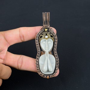 May include: A white carved pendant of a woman with her hands clasped in prayer, wearing a crown with a pearl, set in a copper wire frame.
