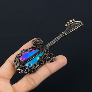 Puede incluir: Un colgante de guitarra envuelto en alambre con una piedra preciosa iridiscente en el cuerpo. La guitarra est&aacute; hecha de alambre de cobre y tiene un dise&ntilde;o detallado. El colgante es una pieza de joyer&iacute;a &uacute;nica y elegante.